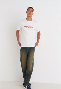 Balta t-krekls ar sarkano "Pepe Jeans" logotipu, apvienots ar gaiši ziliem džinsiem ar izbalējušu apdari. Modelis stāv uz baltā fona.