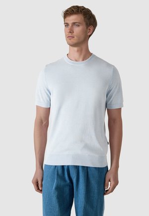 Nuori mies, jolla on lyhyet vaaleanruskeat hiukset, pukeutuneena vaaleansiniseen lyhythihaiseen villapaitaan ja sinisiin housuihin, katsoo sivulle.