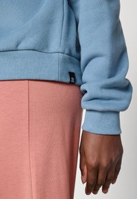Nærbilde av en mørkebrun hånd ved siden av en blå sweatshirterm og rosa bukser, som fremhever teksturer i stoffet og sømmene på plagget.