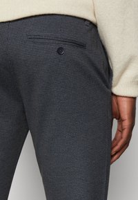 Primo piano di una persona che indossa pantaloni grigio scuro a motivo con una tasca posteriore con bottone e una manica di un maglione beige chiaro visibile.