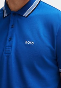 Niebieska koszulka polo z białym kołnierzykiem w paski i haftowanym logo "BOSS" na lewej piersi, noszona przez osobę.