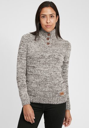 Brun og kremfarget melert strikkegenser med høy krage, tre brune knapper og et tan-farget logo-merke nederst.