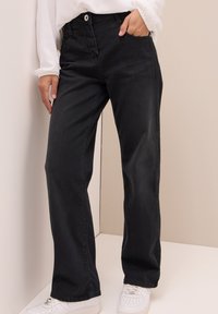 Zwarte denim broek met een recht model, voorzien van voorzakken en een knoopsluiting, gecombineerd met witte sneakers op een effen achtergrond.
