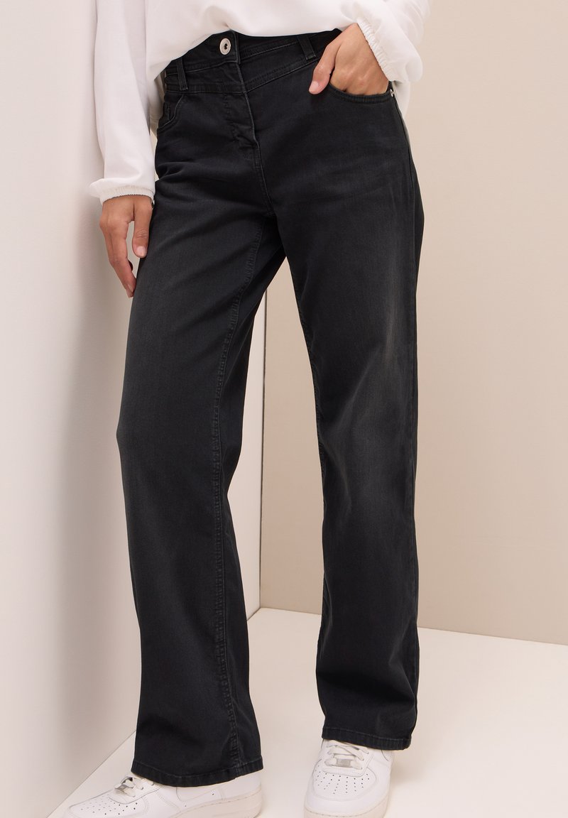 Zwarte denim broek met een recht model, voorzien van voorzakken en een knoopsluiting, gecombineerd met witte sneakers op een effen achtergrond.