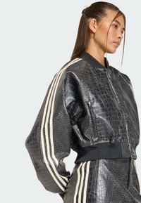 Skraćena crna bomber jakna od umjetne kože s teksturom krokodilske kože, s bijelim prugastim detaljima na rukavima i pripijenim crnim pojasom.