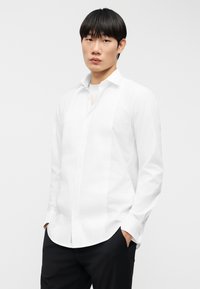 Λευκό πουκάμισο με μακριά μανίκια, με υφή στη ν μπροστινή πλευρά και κλασικό γιακά· φορεμένο πάνω από ανοιχτόχρωμο πουκάμισο, σε συνδυασμό με μαύρο παντελόνι.