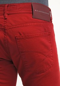 Jeans in denim rosso con una tasca posteriore caratterizzata da un design di cucitura curva distintivo e un'etichetta in pelle con la scritta "BALDESSARINI JEANS" in rilievo.