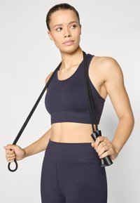 Marineblå sportsbh med ribbet kant, som har en tettsittende design. Modellen holder en svart hoppetau og viser frem den elastiske stoffstrukturen.