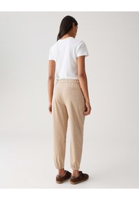 Pantalons de jogging beiges avec des poignets et une taille élastiques, associés à un t-shirt blanc uni. La tenue présente un design minimaliste et une texture lisse.