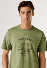 Camisa de algodón verde oliva con mangas cortas, que presenta un gráfico negro de una furgoneta y el texto "LAS COSAS PASAN" arriba.