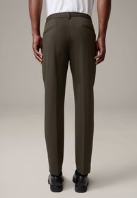 Dunkelolivgrüne, schmal geschnittene Hose aus strukturiertem Stoff, mit elastischem Bund und zwei Gesäßtaschen. Getragen mit schwarzen Schuhen.