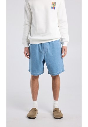Personne portant un short en jean bleu clair, un sweat-shirt blanc à col rond avec un petit motif coloré, des chaussettes blanches et des chaussures beiges sans lacets.