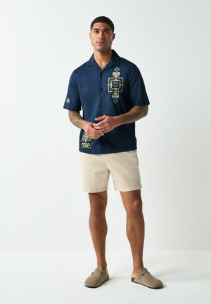 Mand stående iført en marineblå kortærmet skjorte med knapper og geometriske mønstre, beige shorts og taupe slip-on sko, hvid baggrund.