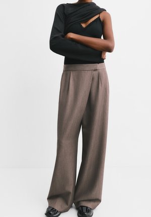 Personne portant un haut noir à une manche, un pantalon large marron ample et des chaussures noires, debout devant un fond blanc uni.