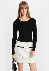 Haut noir à manches longues côtelé associé à une mini-jupe en tweed blanche avec des accents de poches noires. Le mannequin porte un petit sac à main noir.