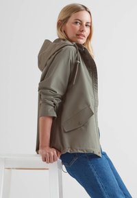 Olive green lightweight jacket with a hood, snap buttons, and two side pockets. Styled with rolled sleeves and paired with blue jeans.