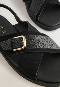 Sandales noires avec une bride texturée noire et une boucle dorée. La tige a une finition en daim, tandis que la semelle est lisse et mate.