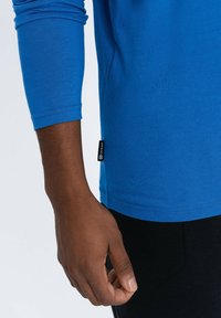 Blauwe lange mouwen shirt met een ronde zoom, met geribde manchetten en een klein label aan de zijkant. Stof lijkt zacht en glad te zijn.