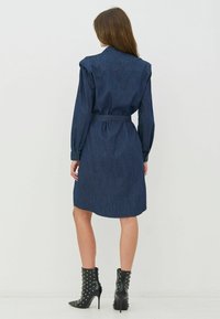 Farmerkék ingetruha, hosszú ujjakkal, gombos mandzsettákkal, öves derékkal, hátul szegélyén kivágásokkal, szegecses fekete bokacsizmával párosítva.