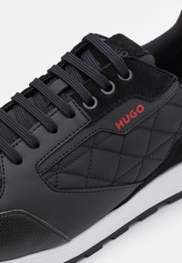 Czarny but sportowy z teksturowaną cholewką z zamszowymi akcentami, pikowanymi wzorami i czerwonym logo. Posiada czarne sznurowadła i białą podeszwę.