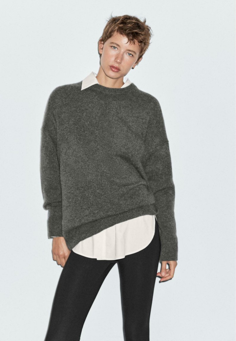 Grå tröja med lös passform och ribbad nederkant, lager på lager över en vit skjorta med krage, matchad med svarta leggings. Mjuk textur, avslappnad design.