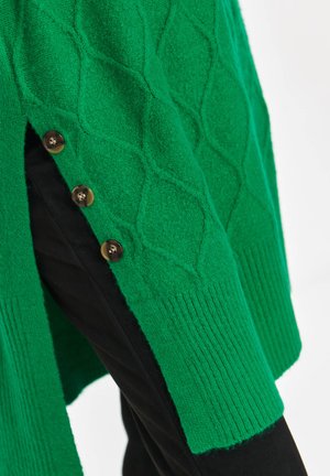 Green knitted sweater with diamond patterns and side slit secured by three brown buttons, worn over black pants.