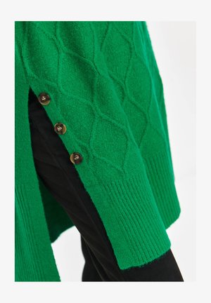 Green knitted sweater with diamond patterns and side slit secured by three brown buttons, worn over black pants.