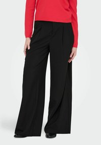 Femme portant un pantalon large noir, un haut à manches longues rouge et des chaussures noires, debout avec une main posée sur la hanche.