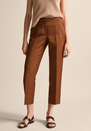 Personne portant un pantalon marron court sur mesure, un haut beige sans manches en maille et des sandales marron foncé, debout devant un fond beige uni.