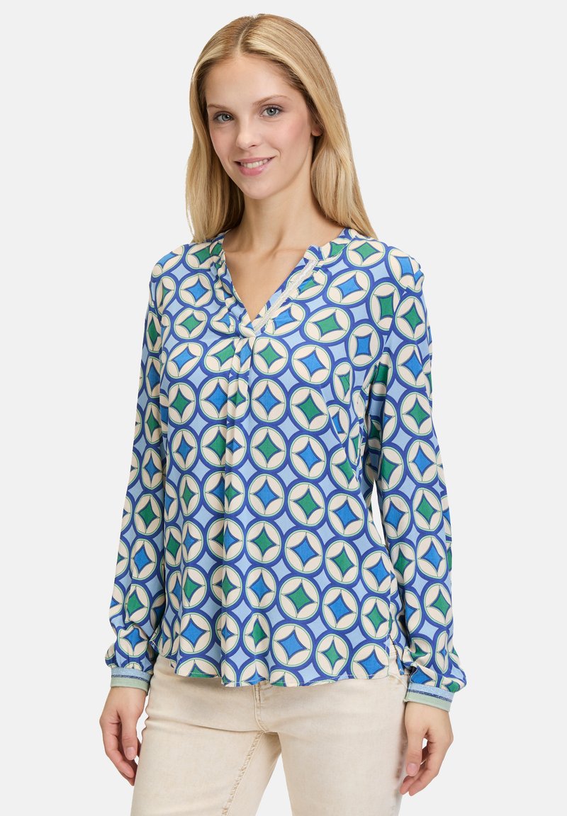 Gemustertes Bluse in Blau mit einem geometrischen Design aus Kreisen und Rauten in Grün und Beige. Lange Ärmel und V-Ausschnitt.