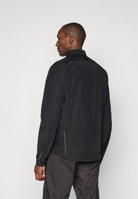 Black lightweight jacket with a high collar, long sleeves, and elastic cuffs. Features side zip pockets and reflective accents for visibility.