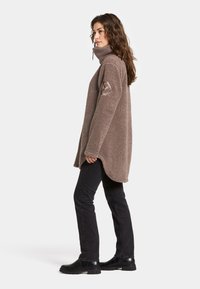 Bruine fleece pullover met hoge kraag, zijsplitten en geborduurd logo op de mouw. Draag het met zwarte broek en laarzen.