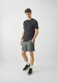 Sort kortærmet skjorte med rund halsudskæring og lomme, kombineret med grå træningsshorts. Modellen har sorte sneakers og hvide sokker på.