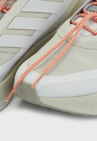Light beige athletic shoes with white stripes, orange and white chevron laces, textured toe, and a rubber sole featuring a logo.