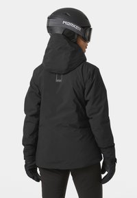Schwarze wasserdichte Skijacke mit Kapuze, strukturiertem Stoff und dezidentem Logo-Detail auf der Rückseite. Kombiniert mit schwarzen Handschuhen für kaltes Wetter.