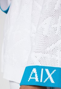 Bílý pletený svetr s texturovanými vzory a modrým lemem. Na rukávu nápis "AIX" v bílé barvě. Měkký, prodyšný materiál.