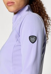 Svetlofialové športové tričko s dlhým rukávom a stojacím golierom, na ľavom rukáve s čiernym logom EA7 a hladkým, elastickým materiálom.