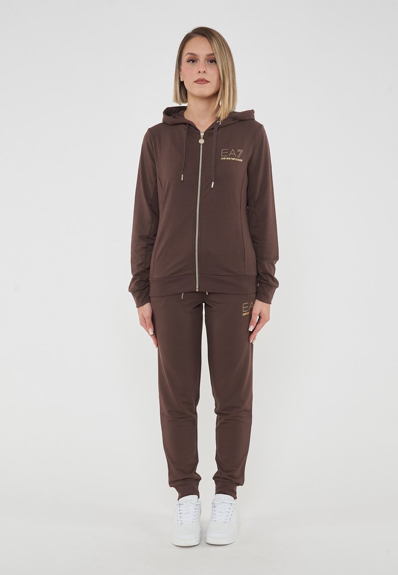 Felpa con zip marrone con logo, pantaloni jogger marroni abbinati e sneakers bianche. Il tessuto sembra liscio con minima texture.