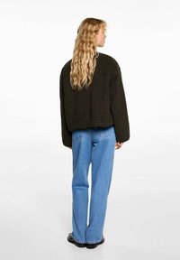 Avskuren, mörkgrön strukturerad jacka med en avslappnad passform, kombinerad med ljusblå högmidjade jeans och svarta skor, sedd från baksidan.