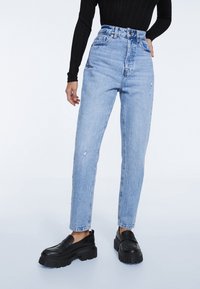 Mujer con jeans de tiro alto, de color azul claro y desgastados, top negro de manga larga con canalé y mocasines de plataforma chunky negros, de pie sobre un fondo blanco.