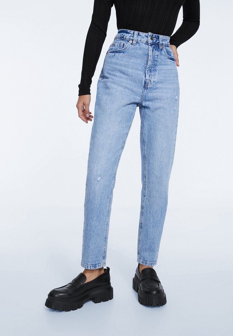 Mujer con jeans de tiro alto, de color azul claro y desgastados, top negro de manga larga con canalé y mocasines de plataforma chunky negros, de pie sobre un fondo blanco.
