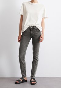 Bílý bavlněný tričko, šedé skinny džíny s worn efektem a černé sandály s přezkou, stojící na světlém pozadí.