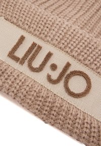 Beige pletená čepice s texturovaným pleteným vzorem. Široký lem s hnědým vyšitým logem "LIU·JO". Měkký a pružný materiál.