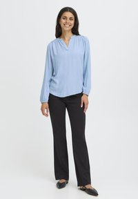 Jeune femme souriante, debout, portant un haut à manches longues bleu clair, un pantalon noir et des chaussures plates noires sur fond blanc.