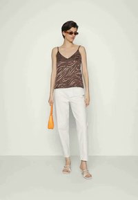 Camisola às riscas em tons de castanho, calças brancas de corte elegante, bolsa laranja e sandálias brancas. Modelo está de pé contra um fundo claro.