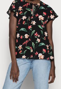 Černá květinová blůza s krátkými rukávy, s vázáním u krku a živým vzorem oranžových a růžových květin s zelenými listy.