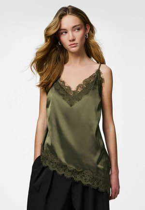 Jeune femme aux longs cheveux portant un caraco en satin vert olive avec une bordure en dentelle et un pantalon noir, debout devant un fond blanc uni.