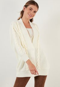 Cardigan de color crema con patrón de trenzas, frontal abierto y tres botones. Presenta mangas largas y puños y dobladillo acanalados.