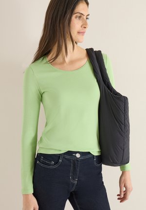 Long-sleeve top in light green fabric with a crew neck. Paired with dark blue jeans featuring contrasting stitching and front pockets. Carrying black shoulder bag.