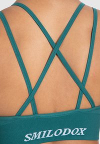 Sujeción deportiva en color teal con un diseño de tirantes en forma de cruz. La tela texturizada presenta el logo "SMILODOX" en blanco en la banda trasera.